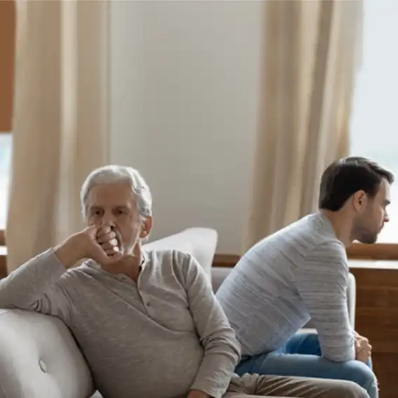 Elderly father and adult son sitting apart on a couch, tension between them